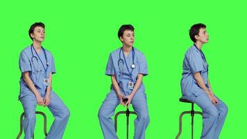 Medical assistant waiting for someone while she sits on chair, feeling impatient and waits for patients to attend checkup appointments. Nurse in scrubs against greenscreen backdrop. Camera A. video