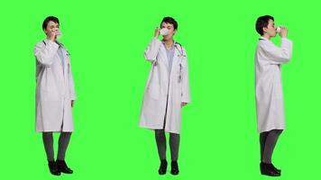 Woman physician drinking a cup of coffee against greenscreen backdrop, taking a break from medical work. Specialist serving caffeine refreshment in studio, wearing a white coat. Camera A. video