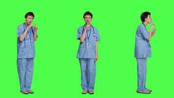 Medical assistant asking to keep quiet and be silent against greenscreen, showing hush mute symbol to keep privacy and silence. Young nurse showing secrecy taboo sign. Camera A. video