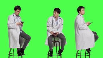 Woman specialist using smartphone social media apps in studio, sitting on a chair against greenscreen backdrop. Physician waiting for someone and browses online webpages, texting. Camera A. video