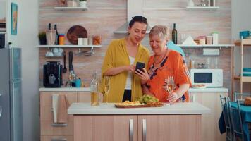 Woman showing a funny movie on smart phone to her mother, sitting in the modern kitchen around the table drinking a glass of white wine. Senior person learning browsing using internet technology video