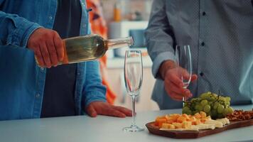 Close up of young man pouring white wine in glasses. Two generations tasting a cup of champagne in cozy dining room while women preparing the healthy dinner video