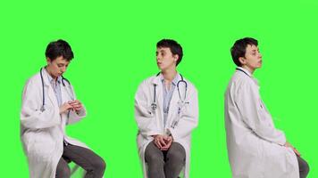 General practitioner acting impatient sitting on a chair against greenscreen backdrop, waiting for patients to attend checkup appointments. Woman specialist in white coat waits for people. Camera B. video