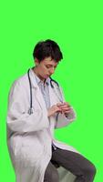 Side view General practitioner acting impatient sitting on a chair against greenscreen backdrop, waiting for patients to attend checkup appointments. Woman specialist in white coat waits for people. Camera B. video