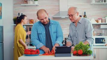 père et fils cuisine des légumes pour dîner en utilisant en ligne recette sur PC ordinateur à Accueil cuisine. Hommes en utilisant numérique tablette tandis que en train de préparer le repas. élargi famille confortable relaxant fin de semaine. video