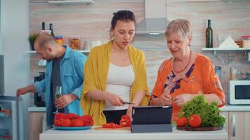 madre y hija corte el pimienta y mirando en tableta, Cocinando utilizando en línea digital tableta receta en ordenador personal computadora a hogar cocina. mientras preparando el comida. extendido familia acogedor relajante fin de semana video