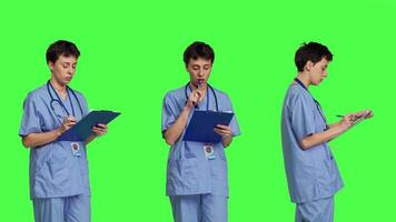 Medical assistant writing checkup information on clipboard files, taking notes and making doctor appointments with checklist. Nurse wearing blue scrubs stands against greenscreen backdrop. Camera B. video