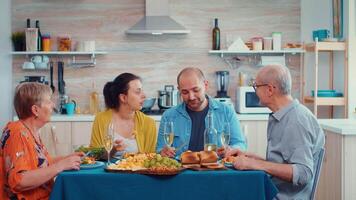 Extended family talking having relaxing time. Multi generation enjoying time at home, in the kitchen sitting by the table, eating dinner together and drinking video