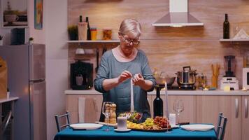 senior vrouw verlichting de kaars aan het wachten haar man voor een romantisch diner. ouderen oud vrouw voorbereidingen treffen feestelijk maaltijd met gezond voedsel voor verjaardag viering, zittend in de buurt de tafel in keuken. video