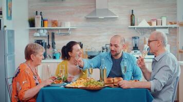 joven hombre hablando durante cena multi generación, cuatro gente, dos contento parejas que se discute y comiendo durante un gastrónomo comida, disfrutando hora a hogar, en el cocina sentado por el mesa. video