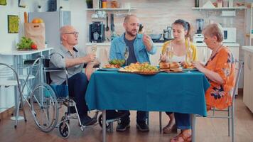 abuelo con invalidez en silla de ruedas y familia teniendo cena. dos contento parejas hablando y comiendo durante un gastrónomo comida, disfrutando hora a hogar sentado alrededor el mesa en el cocina. video