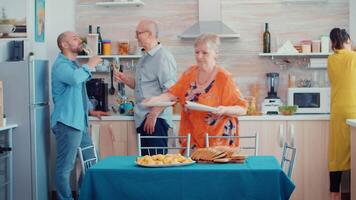 Middle aged woman and older senior have fun working together setting the dinner table in kitchen, while men talking in background and drinking a glass of white wine during a relaxing family day. video