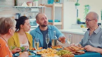 uitgebreid familie interactie terwijl hebben een maaltijd Bij dining tafel in de keuken. multi generatie, vier mensen, twee gelukkig paren pratend en aan het eten gedurende een fijnproever diner, genieten van tijd Bij huis. video