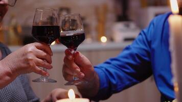 cerca arriba de tintinamente rojo vino lentes durante romántico cena. contento alegre mayor mayor Pareja comida juntos en el acogedor cocina, disfrutando el comida, celebrando su aniversario. video