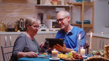 senior gepensioneerd oud man tonen foto's Aan tablet naar zijn vrouw gedurende romantisch diner. paar zittend Bij de tafel, browsen, praten, gebruik makend van internetten, vieren hun verjaardag in dining kamer. video