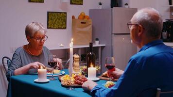 glücklich Senior alt Paar Sitzung beim Essen Tabelle Trinken rot Wein und reden. beim heim, lächelnd Mitte alt reifen Familie genießen romantisch Abendessen und angenehm Konversation auf Mahlzeit zusammen. video