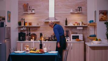 velho idosos homem cozinhando para dele esposa uma romântico jantar, esperando dentro cozinha. Senior velho casal conversando, sentado às a mesa dentro cozinha, desfrutando a refeição a comemorar seus aniversário com saudável Comida video