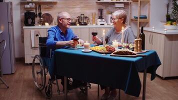 paralysé homme grillage avec du vin avec le sien épouse. Sénior vieux homme dans fauteuil roulant à manger avec vieilli femme séance dans le cuisine. immobilisé paralysé handicapé personnes âgées mari ayant romantique dîner video