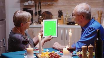 oud gepensioneerd senior paar gebruik makend van groen mock-up scherm digitaal geïsoleerd tablet computer. oud mensen op zoek Bij groen scherm sjabloon chroma sleutel Scherm zittend Bij de tafel in keuken gedurende diner. video