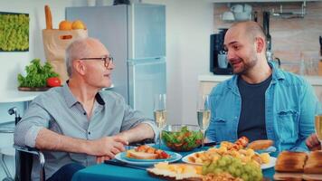 Middle aged man and older senior in wheelchair having fun during family dinner. Happy men smiling, drinking and eating during a gourmet meal, enjoying time sitting around the table in the kitchen video