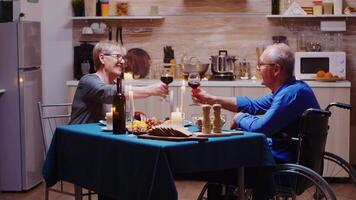 Sénior couple grillage verres à vin séance à le table dans le cuisine. mari dans fauteuil roulant immobilisé paralysé handicapé ayant romantique dîner avec le sien de bonne humeur épouse video