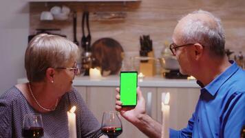 alt Senior im Ruhestand Paar halten Grün Bildschirm Telefon beim Abendessen. alt Menschen suchen beim Attrappe, Lehrmodell, Simulation Vorlage Chroma Schlüssel isoliert Clever Telefon Anzeige mit Technologie Internet Sitzung beim das Tabelle im Küche. video