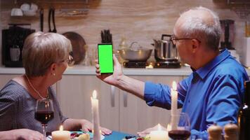 vieux retraité Sénior couple en portant maquette téléphone à dîner. vieilli gens à la recherche à vert écran modèle chrominance clé isolé intelligent téléphone afficher en utilisant technologie l'Internet séance à le table dans cuisine. video