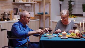 Sénior vieux homme dans fauteuil roulant à manger avec le sien de bonne humeur épouse séance à le table dans le cuisine. immobilisé paralysé handicapé personnes âgées mari ayant romantique dîner video