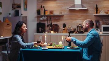 amantes comida juntos, comiendo y Bebiendo vino durante festivo cena en el cocina. contento Pareja hablando, sentado a mesa disfrutando comida a hogar teniendo romántico hora juntos sorpresa vela luces video