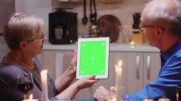 oud gepensioneerd senior paar gebruik makend van groen mock-up scherm digitaal geïsoleerd tablet computer. oud mensen op zoek Bij groen scherm sjabloon chroma sleutel Scherm zittend Bij de tafel in keuken gedurende diner. video