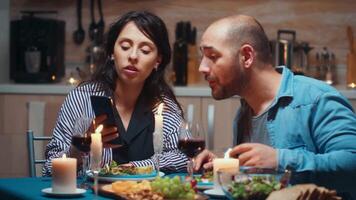 Young wife showing photos on smartphone to her husband during romantic dinner. Couple sitting at table talking using phone browsing searching internet technology surfing online celebrating anniversary video