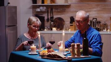 Porträt von Senior Paar mit rot Wein Brille Sitzung beim das Tabelle im das gemütlich Küche. glücklich heiter Alten Paar Essen zusammen beim heim, genießen das Mahlzeit, feiern ihr Jahrestag video