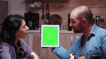 Cheerful young couple using green mock-up screen digital isolated tablet computer. Husband and wife looking at green screen template chroma key display sitting at the table in kitchen during dinner. video