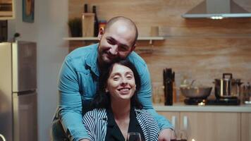 portret di allegro coppia guardare a telecamera, sorridente, seduta a tavolo nel cucina durante romantico cena. contento sposato gente speciale tenero momenti, divertirsi il pasto a candela luci celebrazione video