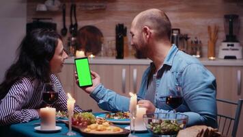 Jeune content couple en portant vert écran téléphone à dîner. de bonne humeur gens à la recherche à maquette modèle chrominance clé isolé intelligent téléphone afficher en utilisant technologie l'Internet séance à le table dans cuisine. video