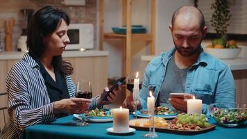 Focused married couple reading news of phones during romantic dinner. Adults sitting at the table, browsing online, surfing, using smartphones, celebrating their anniversary in the dining room. video