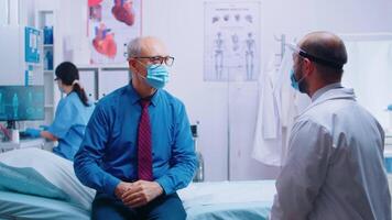 Senior man at doctor appointment during COVID-19 pandemic. Patient wearing mask and doctor in protective wear. Healthcare consultation, medicinal system. Private modern clinic video