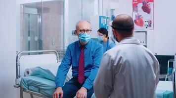 dokter ziekenhuis medisch overleg gedurende covid-19 globaal pandemie. oud gepensioneerd senior Mens vervelend een masker en gezondheidszorg arbeider in beschermend uitrusting voor overleg. modern privaat kliniek video