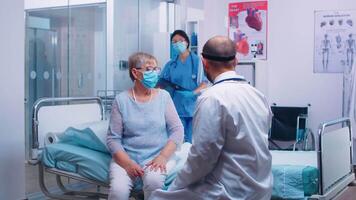 Medical stuff in visor explainingX Ray result to old retired woman sitting on hospital bed during COVID-19 pandemic. Nurse checking with old man in background. Modern private clinic or healthcare facility video