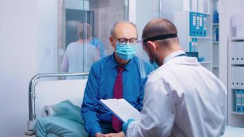 läkare läsning medicinsk resultat medan bär skyddande mask och de patient är Sammanträde på sjukhus säng. modern privat sjukhus eller klinik. sjukvård medicinsk läkare samråd under covid-19 pandemi video