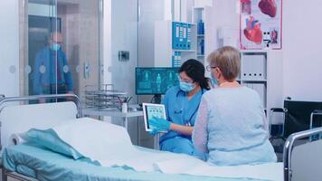 Nurse and doctor with protective equipment and wearing masks offering medical consultation during coronavirus outbreak in modern clinic. Patient sitting on hospital bed looking at tablet PC video