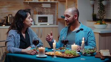 Jeune marié couple profiter romantique dîner grillage et en buvant rouge du vin séance à le table dans confortable cuisine. content adultes à manger ensemble le repas célébrer leur anniversaire à bougie lumières video