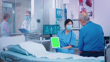 Nurse presenting green screen tablet to patient in modern private hospital. Isolated mockup chroma replacement screen on gadget for your app, text, video or digital assets. Easy keying medicine medical related theme