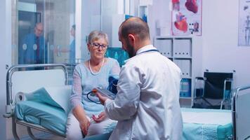 Physician measuring blood pressure with a digital tonometer to senior old retired woman in modern private clinic while sitting in hospital bed. Medical medicine healthcare system disease treatment video