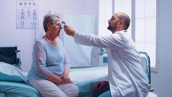 médico medición temperatura a mayor mujer quien se sienta en hospital cama. utilizando digital infrarrojo sin contacto termómetro en moderno privado clínica. médico examen para infecciones y enfermedad video