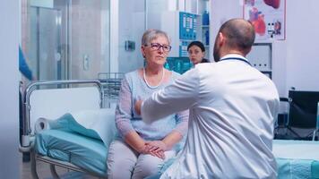 Doctor measuring old senior woman temperature with digital infrared contactless thermometer in modern private clinic. Patient sitting on hospital bed, healthcare system ilness and disease prevention video