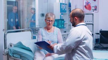 Senior patient signing medical decisions form sitting on hospital bed in modern private clinic. Doctor with clipboard, nurse working in backgorund. Healthcare medical medicinal system documents contract video