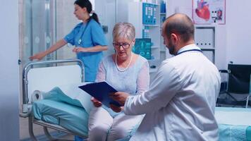 Senior retired woman signing for medical release in modern private clinic sitting on hospital bed, nurse working in background. Healthcare medicine consultation doctor patient video