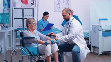 Doctor helping disabled senior patient to recover muscle strength in private modern rehabilitation clinic or hospital. Nurse in background talking with elderly man with walking frame video