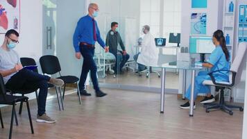 Senior man discussing with nurse at hospital reception wearing mask against coronavirus, assistant with sterile gloves is pushing disabled mature woman in wheelchair through waiting area. video